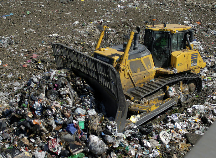 A yellow bulldozer atop a mound of trash, moving debris at a landfill.